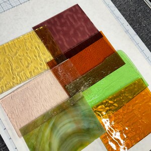May include: Assortment of glass sheets in various colors and textures, including yellow, orange, green, brown, and white. The glass sheets are arranged on a white surface.