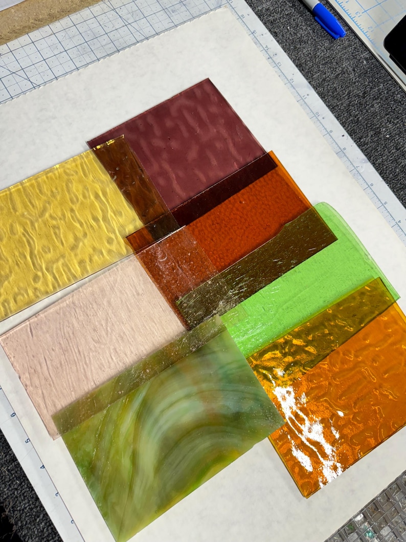 May include: Assortment of glass sheets in various colors and textures, including yellow, orange, green, brown, and white. The glass sheets are arranged on a white surface.