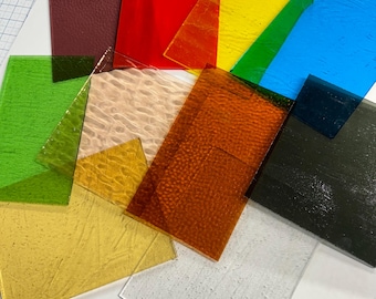 May include: Assortment of textured glass sheets in various colors, including red, yellow, green, blue, brown, and white. The sheets are arranged in a random pattern on a white surface.