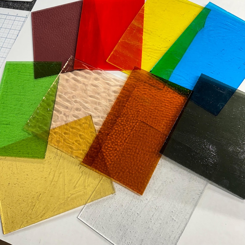 May include: Assortment of textured glass sheets in various colors, including red, yellow, green, blue, brown, and white. The sheets are arranged in a random pattern on a white surface.