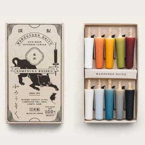 Puede incluir: Un juego de diez velas cilíndricas de salvado de arroz en una caja de cartón. La tapa muestra una ilustración de un gato negro, el texto Warousoku Daiyo, Komenuka Rosoku y 100% Quality. Las velas están dispuestas en dos filas.