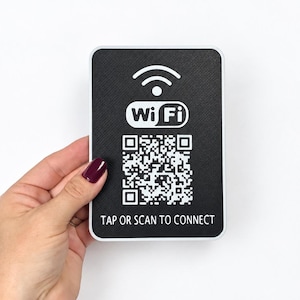 May include: A rectangular black sign with a white border featuring a white Wi-Fi symbol, the text "Wi Fi", a central QR code, and the text "TAP OR SCAN TO CONNECT" printed in white at the bottom of the sign surface.