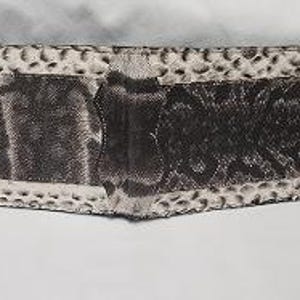May include: A bi-fold wallet featuring a textured, snakeskin-patterned exterior in shades of black, grey, and off-white. The wallet has a horizontal orientation with a central seam and a repeating scale-like pattern across the entire surface.