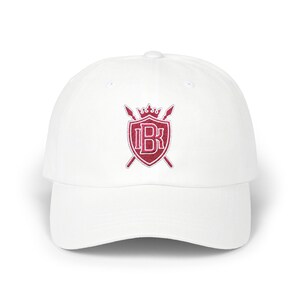 May include: A white baseball cap featuring an embroidered design on the front. The emblem consists of a red shield with the letters IBK in white, topped with a crown and positioned over two crossed spears in red with white outlines.