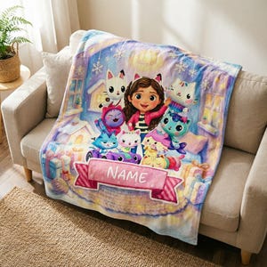May include: A soft throw blanket featuring a colourful illustration of a girl with brown hair surrounded by various cartoon cats in a winter setting with snowflakes and gift boxes. A pink banner at the bottom contains the white text NAME.
