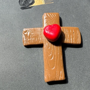 May include: A brown cross with a wood grain texture and a smooth, glossy red heart attached to the center. Above the cross is a gold-colored graphic of a sun rising over a dark, textured circular shape, all set against a dark gray background.