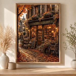 Może przedstawiać: Oprawiony wydruk ścienny przedstawiający brukowaną ulicę jesienią z księgarnią o nazwie The Old Bookshop. Witryna sklepowa posiada duże okna wypełnione książkami, rower zaparkowany na zewnątrz oraz złote liście otaczające budynek.