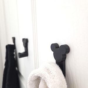 May include: A set of matte black wall-mounted hooks, each featuring a distinct silhouette shaped like a circular head with two rounded ears. These functional hooks are attached to a white surface, with one holding a white towel and another a black towel.