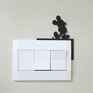 Peut inclure: Une plaque d'interrupteur triple blanche avec une silhouette noire d'un personnage de souris de dessin animé debout sur une petite étagère fixée au coin supérieur droit du couvercle rectangulaire, comprenant trois interrupteurs à bascule.