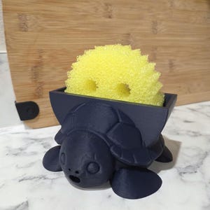 May include: A dark navy blue turtle-shaped sponge holder with a textured matte finish. The turtle features a shell that forms a rectangular container, currently holding a bright yellow, round, porous cleaning sponge with two visible holes.