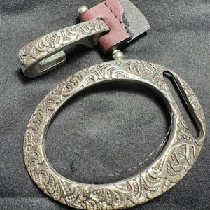 May include: A silver-toned metal oval ring with an intricate embossed pattern, attached to a dark maroon leather strap and a matching silver-toned metal hook clasp. The metal surfaces feature a textured, repeating decorative design throughout.