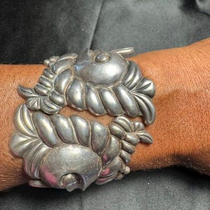 May include: A silver cuff bracelet featuring two mirrored, stylized shell-like motifs with curved, ribbed segments and a smooth, rounded center. The metal has a polished finish with visible oxidation in the recessed areas, creating a high-contrast look.