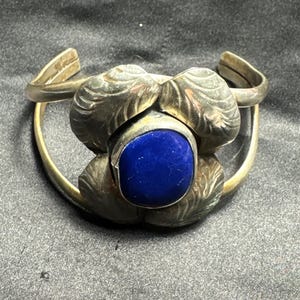 Puede incluir: Un brazalete de plata con una piedra central de lapislázuli azul profundo y lisa. La piedra está rodeada por cuatro pétalos grandes de plata texturizada con detalles de vetas grabadas. El brazalete tiene un diseño de banda abierta.