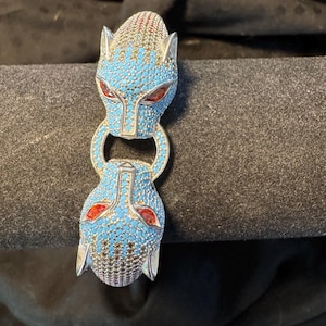May include: A hinged bracelet featuring two panther heads facing each other, connected by a circular ring. The panther heads are encrusted with small blue stones, accented with red stones for the eyes and metallic silver-toned detailing.