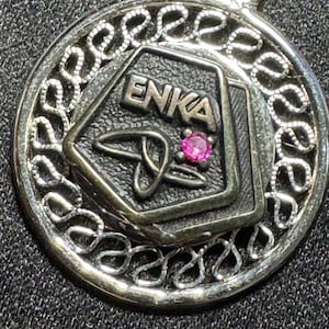 May include: A circular silver-toned pendant featuring an intricate filigree border. The center contains a pentagonal shape with the embossed text ENKA, a stylized linear symbol, and a single small, round, vibrant pink gemstone set in the metal.
