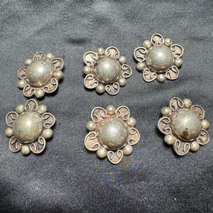 May include: A set of five circular silver-toned metal buttons, each featuring a smooth, raised central dome surrounded by a decorative border of small metal spheres and intricate, swirling wire-like filigree patterns arranged in a floral shape.