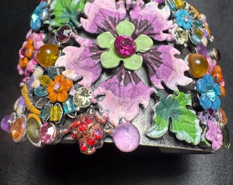 May include: A wide cuff bracelet featuring a dark metal base adorned with colorful enamel flowers in shades of pink, green, orange, and blue. The design is embellished with various faceted rhinestones, cabochons, and small ladybug figures.