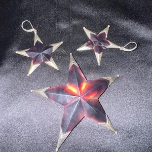 Puede incluir: Un conjunto de tres objetos en forma de estrella con centros de color marrón rojizo oscuro y translúcido, con bordes metálicos plateados. Dos de las estrellas son más pequeñas e incluyen ganchos para las orejas, mientras la tercera es mayor.