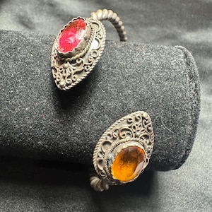 May include: A silver cuff bracelet featuring two teardrop-shaped settings with intricate filigree metalwork. One setting holds a faceted oval red stone, while the other holds a faceted oval amber-colored stone. The band has a twisted rope texture.