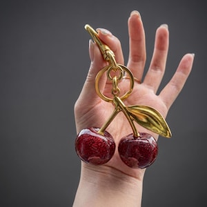 Puede incluir: Un llavero de metal dorado con dos dijes de cerezas redondas, de color rojo oscuro y con purpurina, unidos a una anilla central. Las cerezas están conectadas por un tallo y una hoja dorados, con un cierre de mosquetón resistente en la parte superior.