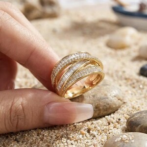 Puede incluir: Un anillo de oro con un diseño cruzado de varias bandas. La superficie está incrustada con numerosas piedras pequeñas y claras, con tallas baguette y redonda, que cubren todo el exterior visible de la banda metálica en un patrón denso y brillante.