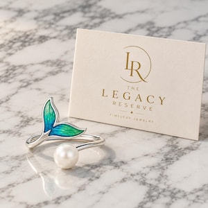 Puede incluir: Un anillo abierto de tono plateado con una perla blanca en un extremo y una cola de sirena esmaltada en azul y verde en el otro, junto a una tarjeta con el texto dorado LR The Legacy Reserve Timeless Jewelry.
