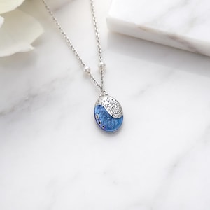 Op de afbeelding: Een zilverkleurige ketting met een fijne schakelketting, twee kleine witte kralen en een ovaal hangertje. De hanger heeft een bovenkant met gedetailleerd zilverfiligraan en een onderkant met een levendig blauw, wervelend iriserend patroon.