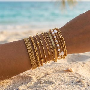 Puede incluir: Un conjunto de nueve pulseras doradas con diversos diseños, que incluyen una banda de malla ancha, una cadena de cuerda trenzada, una cadena fina con una pequeña cuenta, una cadena de eslabones de barra, una cadena de eslabones rectangulares, una cadena plana y una hilera de perlas.