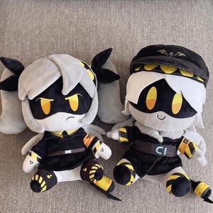 May include: Two plush dolls featuring black and white color schemes. The left doll has grey hair in pigtails and yellow eyes. The right doll wears a black cap with a yellow band, white hair, and yellow eyes. Both wear black jackets with yellow accents.
