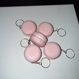 May include: A collection of five light pink macaron-shaped keychains, each featuring a silver-toned metal ring and chain. The round, smooth-textured macarons have a visible white filling layer between the two halves, arranged in a cluster.