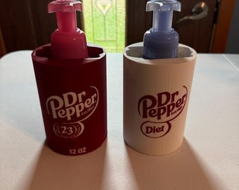 Może przedstawiać: Dwa cylindryczne dozowniki do mydła z pompkami. Lewy dozownik jest bordowy z logo Dr Pepper, liczbą 23 i napisem 12 oz. Prawy dozownik jest biały z logo Dr Pepper i słowem Diet. Oba posiadają różowe i niebieskie pompki dozujące.