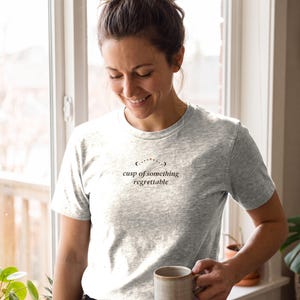 May include: A heather gray short-sleeved t-shirt featuring a crew neckline and black text printed on the chest that reads "cusp of something regrettable" positioned beneath a small arc of dots and a single star.
