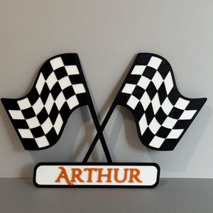 May include: A decorative sign featuring two crossed black and white chequered racing flags positioned above a rectangular white base. The name ARTHUR is displayed in bold orange capital letters on the front of the base, centred horizontally.