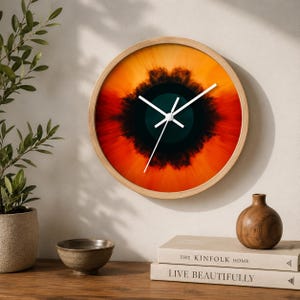 Op de afbeelding: Een ronde wandklok met een lichthouten lijst en een levendige abstracte wijzerplaat met een donkergroenblauw centrum, omringd door een stralend patroon van dieprode en feloranje tinten, voorzien van eenvoudige witte wijzers.