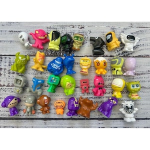 May include: A collection of small, colorful plastic monster figurines in various shapes and expressions. The set includes diverse characters in shades of red, yellow, green, blue, purple, black, and white, each featuring unique facial features.