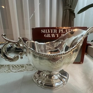May include: A silver-plated gravy boat with a polished metallic finish, featuring an ornate, curved handle and a decorative rope-style rim. The vessel sits on a pedestal base and is positioned in front of a box labeled SILVER PLATED GRAVY BOAT.