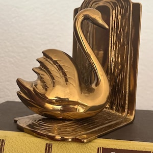 May include: A polished brass bookend featuring a three-dimensional swan figure attached to an L-shaped base. The swan has a curved neck and detailed wing feathers. The base and the vertical back panel have a textured, rippled surface finish.