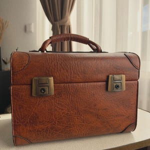 May include: A rectangular brown suitcase with a textured, leather-like finish, featuring a sturdy top handle, reinforced corner detailing, and two antique brass-toned metal latches on the front for secure closure and a structured, boxy silhouette.
