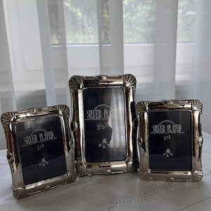 May include: Three silver-plated picture frames with ornate, scalloped corners. The frames contain dark blue inserts with the text "SILVER PLATED" and dimensions of 5.08x7.62 cm for the two smaller frames and 8.89x12.7 cm for the larger centre frame.