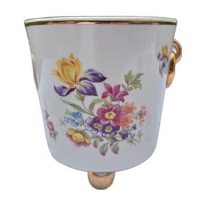 Może przedstawiać: Białe porcelanowe wiaderko z kolorowym wzorem kwiatowym w odcieniach żółci, fioletu, różu i pomarańczy. Naczynie posiada złocony brzeg, złoty uchwyt z boku oraz wspiera się na złotej podstawie w formie stopki.