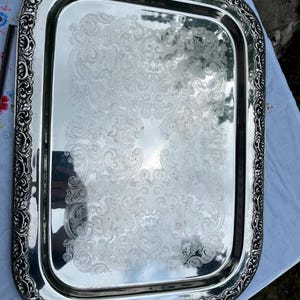 Può includere: Un vassoio da portata rettangolare color argento con una superficie lucida altamente riflettente. Il centro del vassoio è decorato con un intricato motivo a volute inciso, mentre l'ampio bordo presenta un rilievo floreale ornamentato.