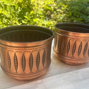 Può includere: Due vasi rotondi in rame caratterizzati da un motivo in rilievo ripetuto di forme ovali verticali con linee tratteggiate centrali. I contenitori metallici presentano una calda finitura color marrone rossiccio e un interno scuro.