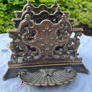 May include: A dark bronze-toned metal letter holder featuring an intricate openwork design with scrollwork and floral motifs. The piece has three vertical dividers and a scalloped, shell-shaped base tray at the front for holding small items.