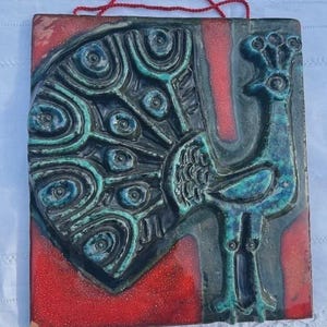 Op de afbeelding: Een vierkante keramische wandplaat met een reliëf van een pauw in tinten blauw en groenblauw. De achtergrond is felrood met een gespikkelde textuur. Aan de bovenkant is een dun rood touwtje bevestigd om het object op te hangen.