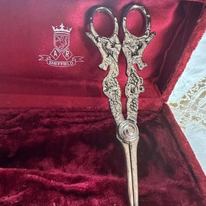 May include: A pair of silver-toned metal grape shears with ornate embossed handles featuring floral and vine motifs, resting inside a rectangular case lined with deep red velvet. The interior lid displays a white logo with the text A R SHEFFIELD.