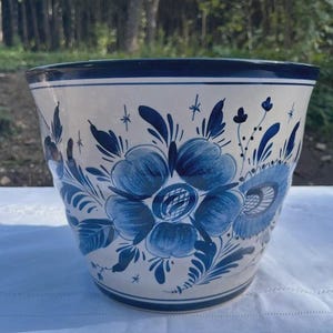 Könnte beinhalten: Ein Keramik-Pflanzgefäß mit cremefarbener Basis und handgemalten dunkelblauen Blumenmotiven sowie Blattwerk. Das Design zeigt große zentrale Blüten, kleine Knospen und sternförmige Akzente, abgerundet durch einen dunkelblauen Rand und Boden.