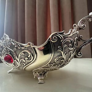 Op de afbeelding: Een zilverkleurige decoratieve schaal met een sierlijk, verhoogd krulpatroon aan de buitenkant. De schaal rust op drie kleine pootjes en bevat een opvallende, gladde, ronde rode edelsteen die aan de zijkant nabij de rand is geplaatst.
