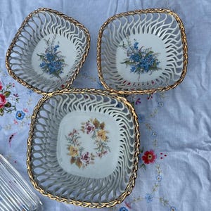 Pode incluir: Três taças quadradas de porcelana com lados de treliça aberta e bordas douradas. Duas taças apresentam desenhos florais azuis no centro, enquanto a terceira exibe uma coroa central de rosas amarelas e cor-de-rosa. Um prato de vidro é visível.