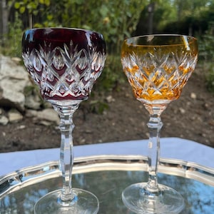 Puede incluir: Dos copas de vino de cristal con un patrón de corte de diamante. Una copa presenta un cuenco de color rojo intenso, mientras que la otra tiene un cuenco de color ámbar. Ambas copas tienen tallos transparentes facetados y bases circulares.