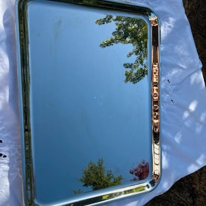 Op de afbeelding: Een rechthoekig zilverkleurig metalen dienblad met een gepolijste, spiegelende afwerking. Het dienblad heeft afgeronde hoeken en een verhoogde, gladde rand. Het oppervlak reflecteert de buitenomgeving, inclusief bomen en de blauwe lucht.